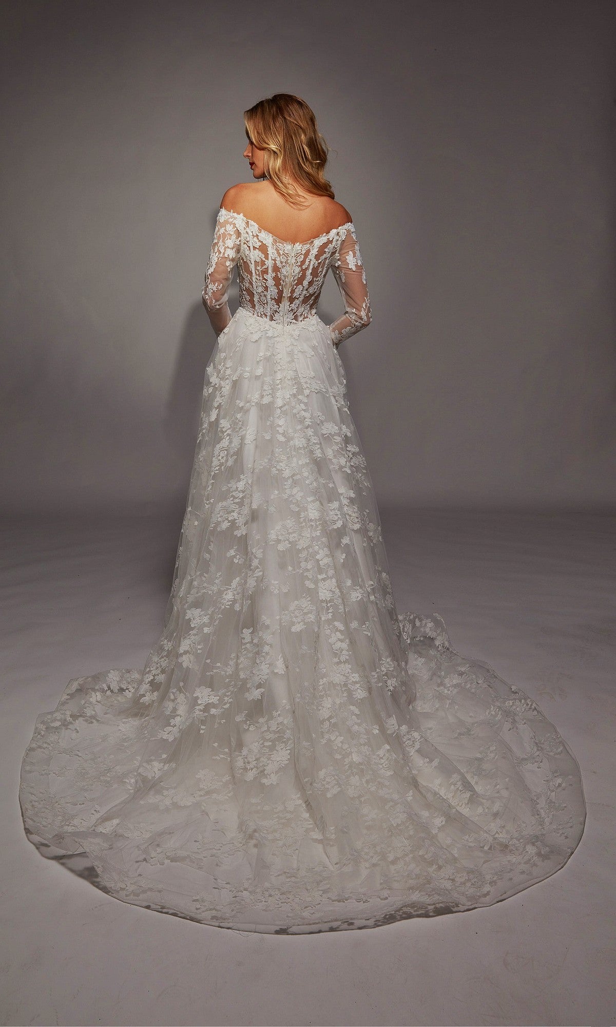 A woman faces away, wearing the Alyce 7150: a long white off-the-shoulder lace wedding dress with long sleeves, a flowing train, and delicate ruffle details.
